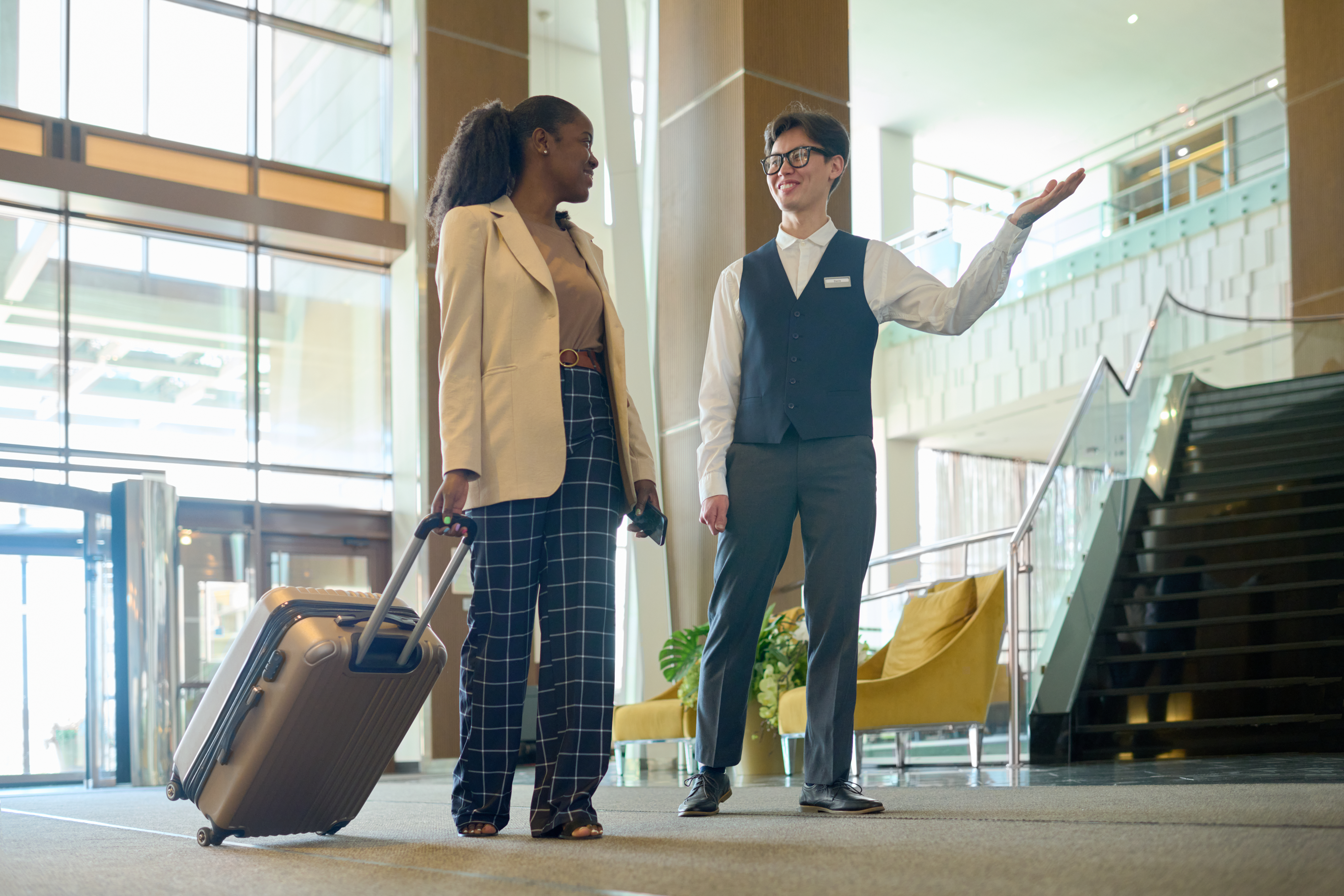 Young smiling manager of modern hotel consulting female traveler with suitcase entering lounge and moving towards reception counter